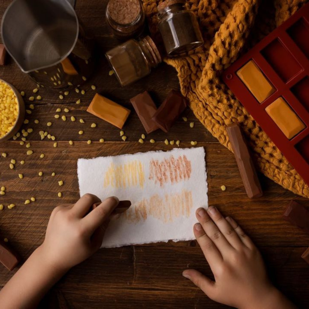 Person's hands using handmade, natural crayons to draw on a white peice of paper surrounded by ingredients used to make them including beeswax pellets and pigments.