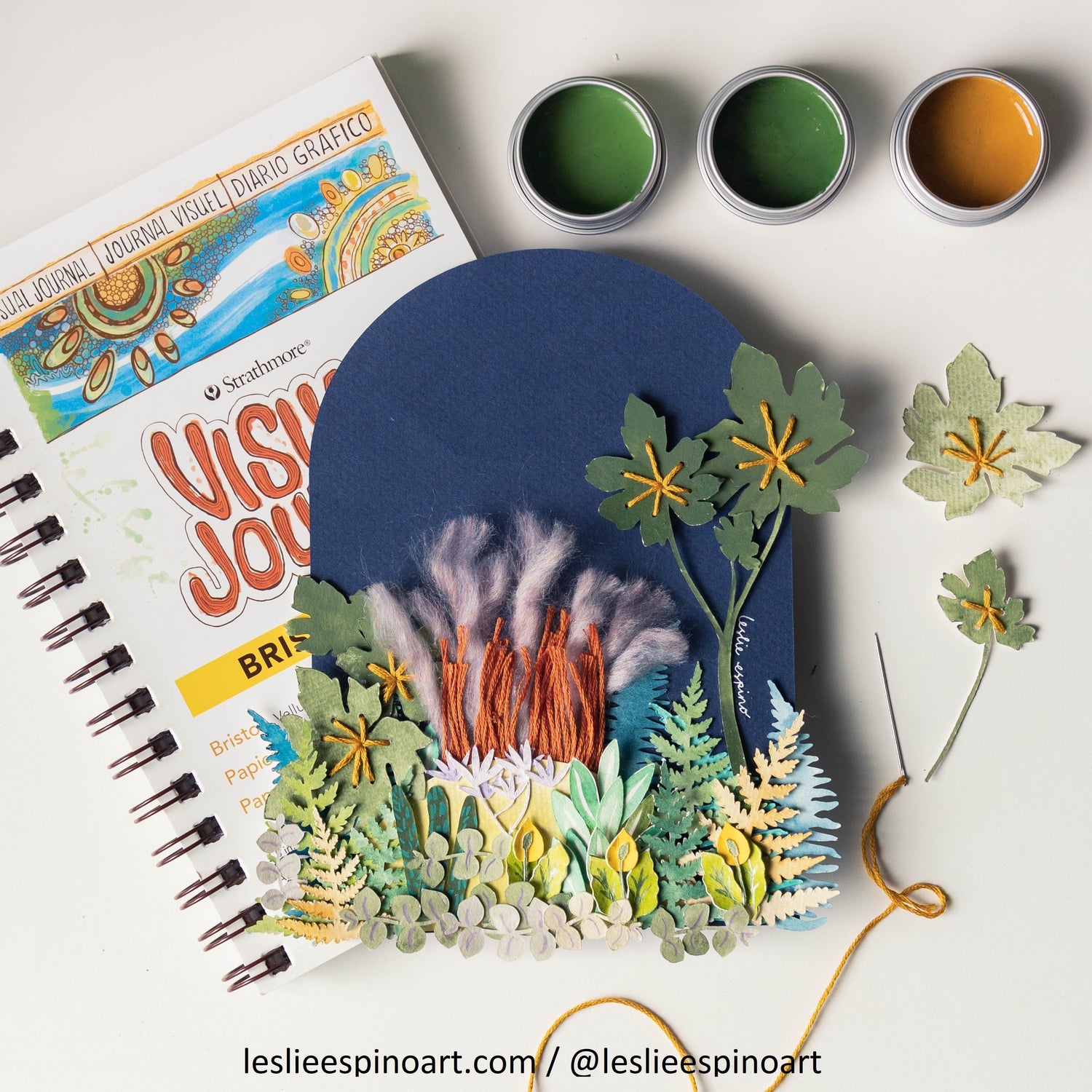 One of Leslie Espino's botanical paper and thread creation in front of their book, "Visual Journal" with 3 containers in the upper right hand corner containing light green, dark green and yelllow ochre paint.
