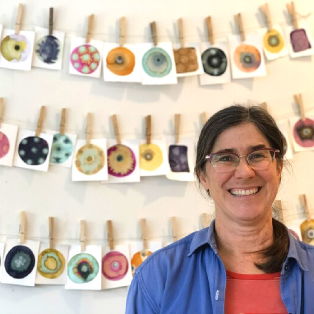 Printmaker Marjorie Morgan standing in front of a wall with 3 rows of small works of art hung up by clothes pins.