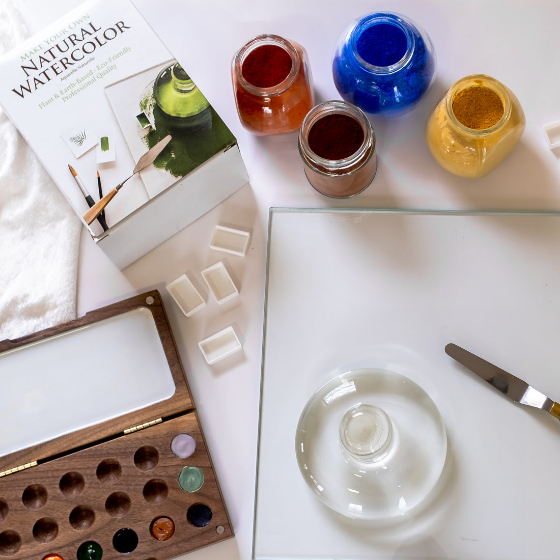 A glass palette, palette knife and glass muller next to four open jars of paint, four empty watercolor pans, a wooden watercolor palette and a Natural Watercolor Kit