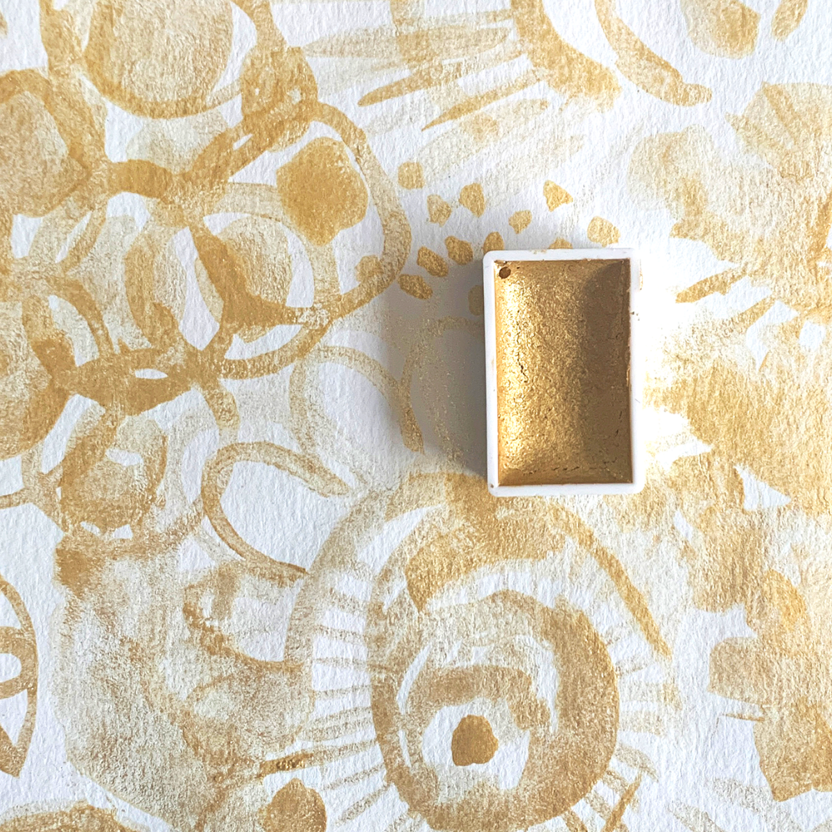 A pan of handmade gold paint made by Kristen Fagan sitting on a white background with gold paint strokes.