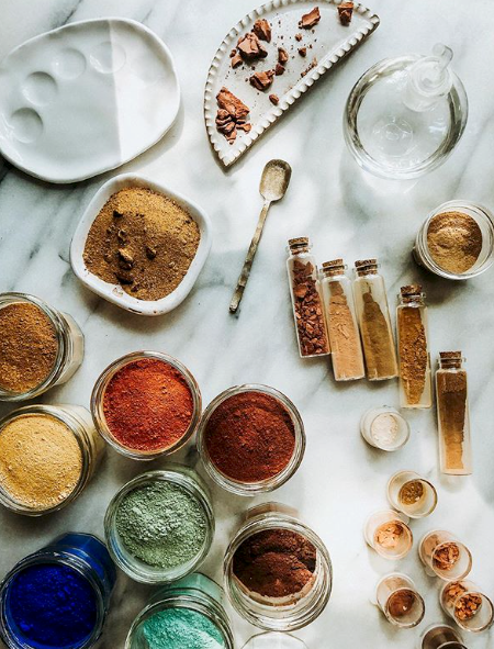 Overhead view of a white marble table covered with a rainbow of natural pigments in glass jars and glass viles, next to a paint palette, glass muller and palette knife