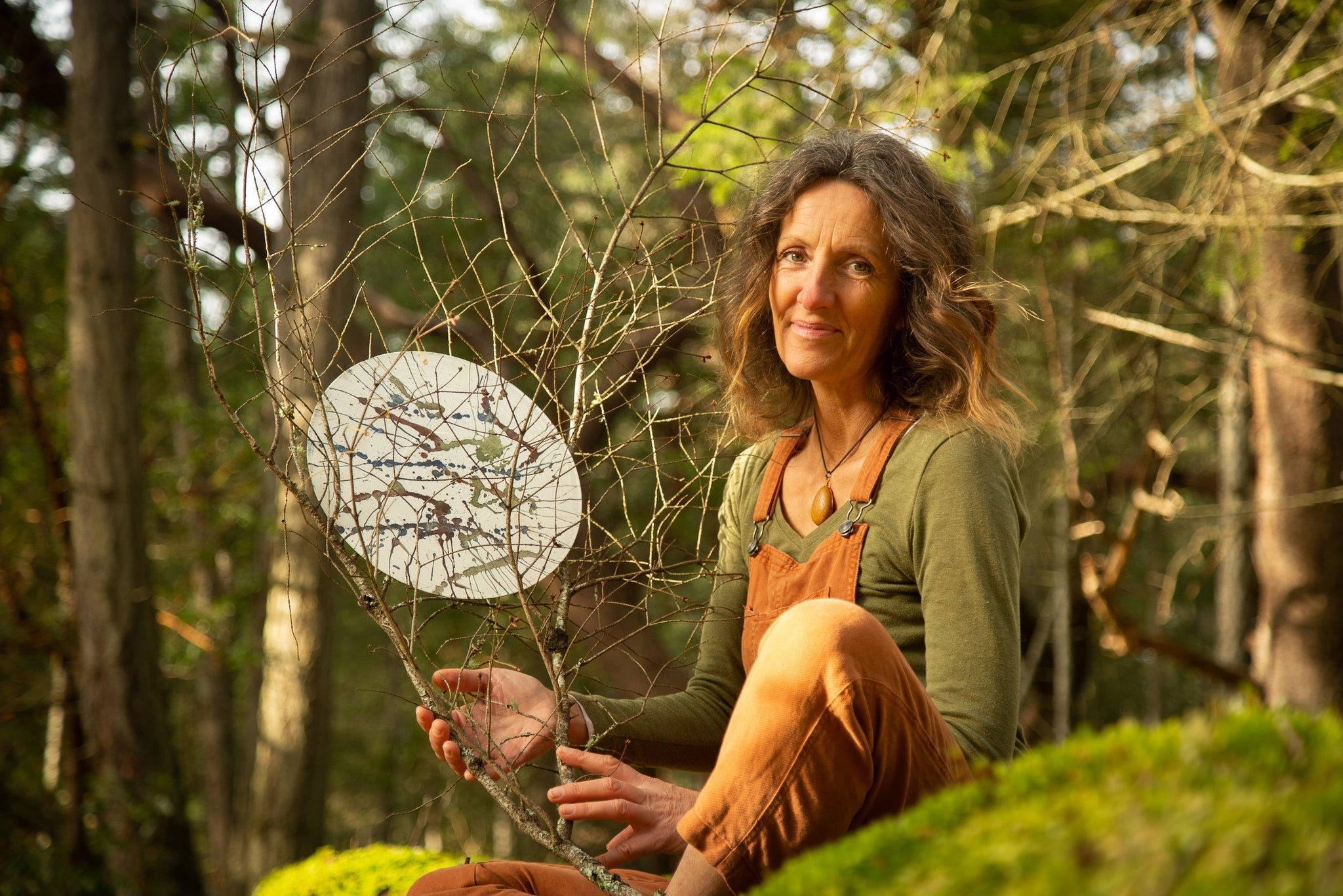 Artist Marghanita Hughes sitting in the woods on a mossy rock holding a stick sculpture.