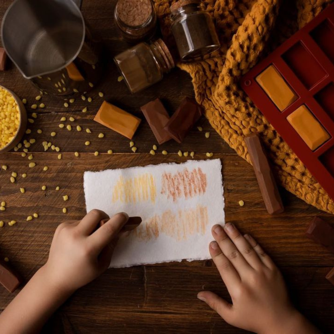 Person's hands using handmade, natural crayons to draw on a white peice of paper surrounded by ingredients used to make them including beeswax pellets and pigments. 