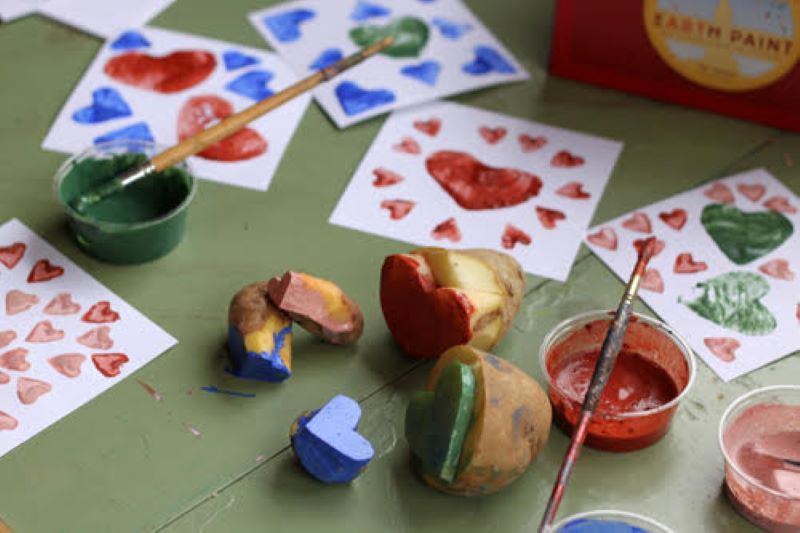 Table filled with cards made from potato stamps sitting next to the potatoes with paint containers of red, green and blue paint with paint brushes 