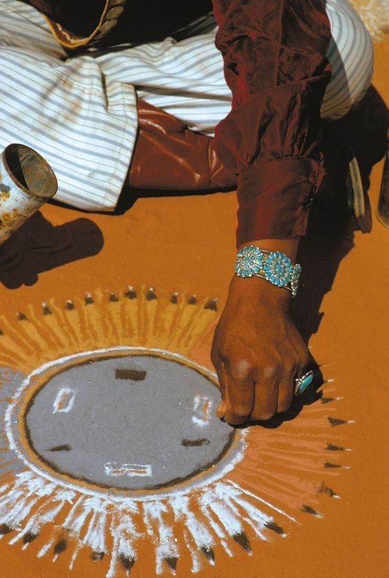 Native American person sitting on the ground and using their hand to carefully sprinkle pigment into an image