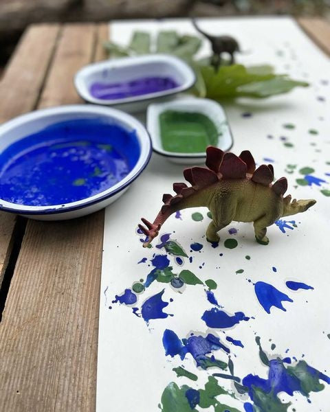 Photo of a small toy dinosaur on top of a piece of white paper that has been decorated with blue, green and purple paint, sitting next to two dishes containing the paint