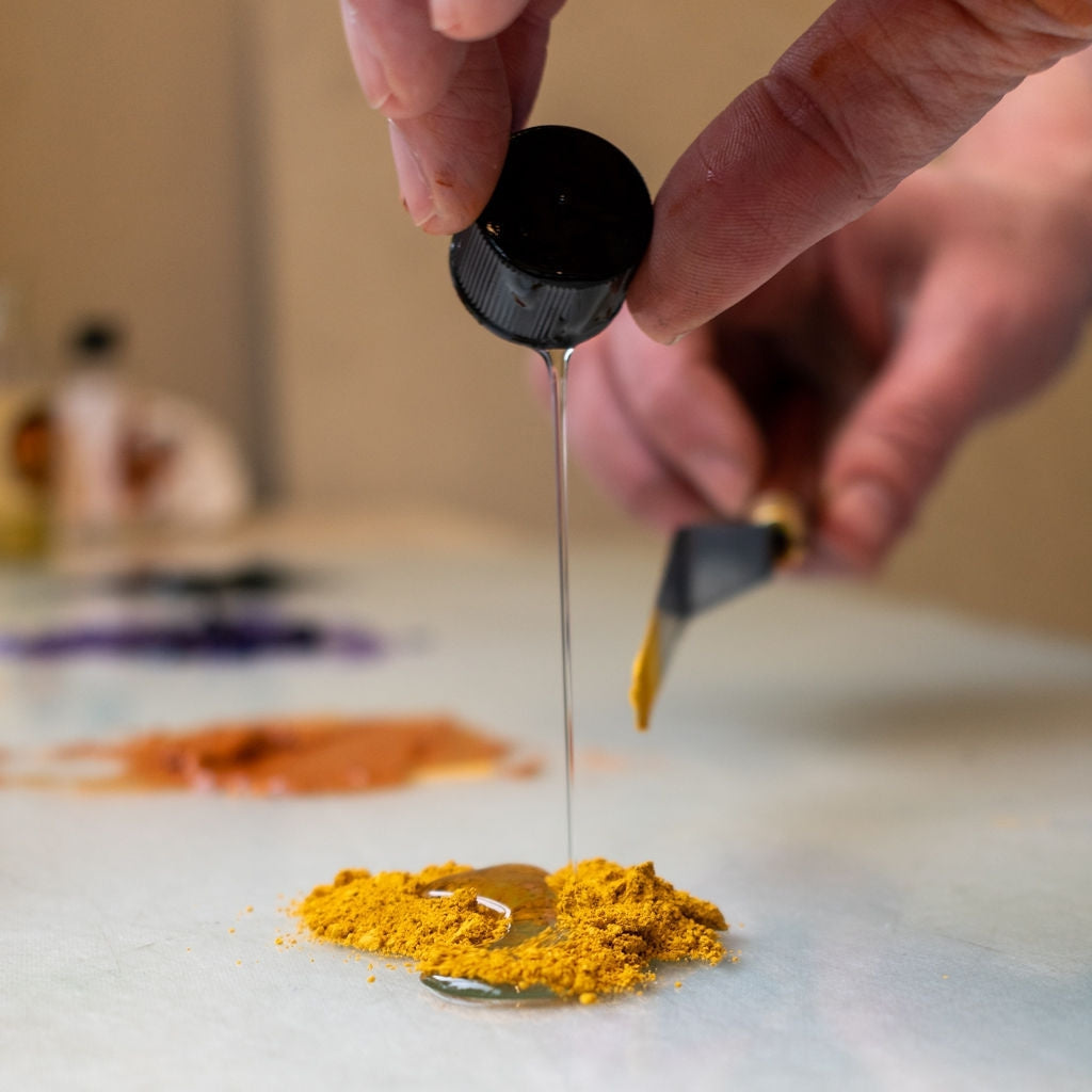 Person's hand pouring walnut ink into a pile of yellow Earth & Mineral Pigment with a palette knife in their other hand