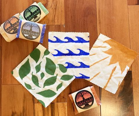 3 hemp flags painted with (left to right) green leaves, blue waves and a yellow sun surrounded by 4 Natural Earth Paint packets