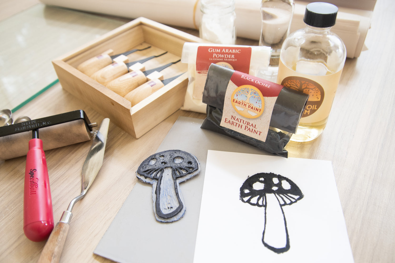 Print of a black mushroom on white paper made with ink made from natural Earth & Mineral Pigments and walnut oil surrounded by carving tools, the block and a brayer