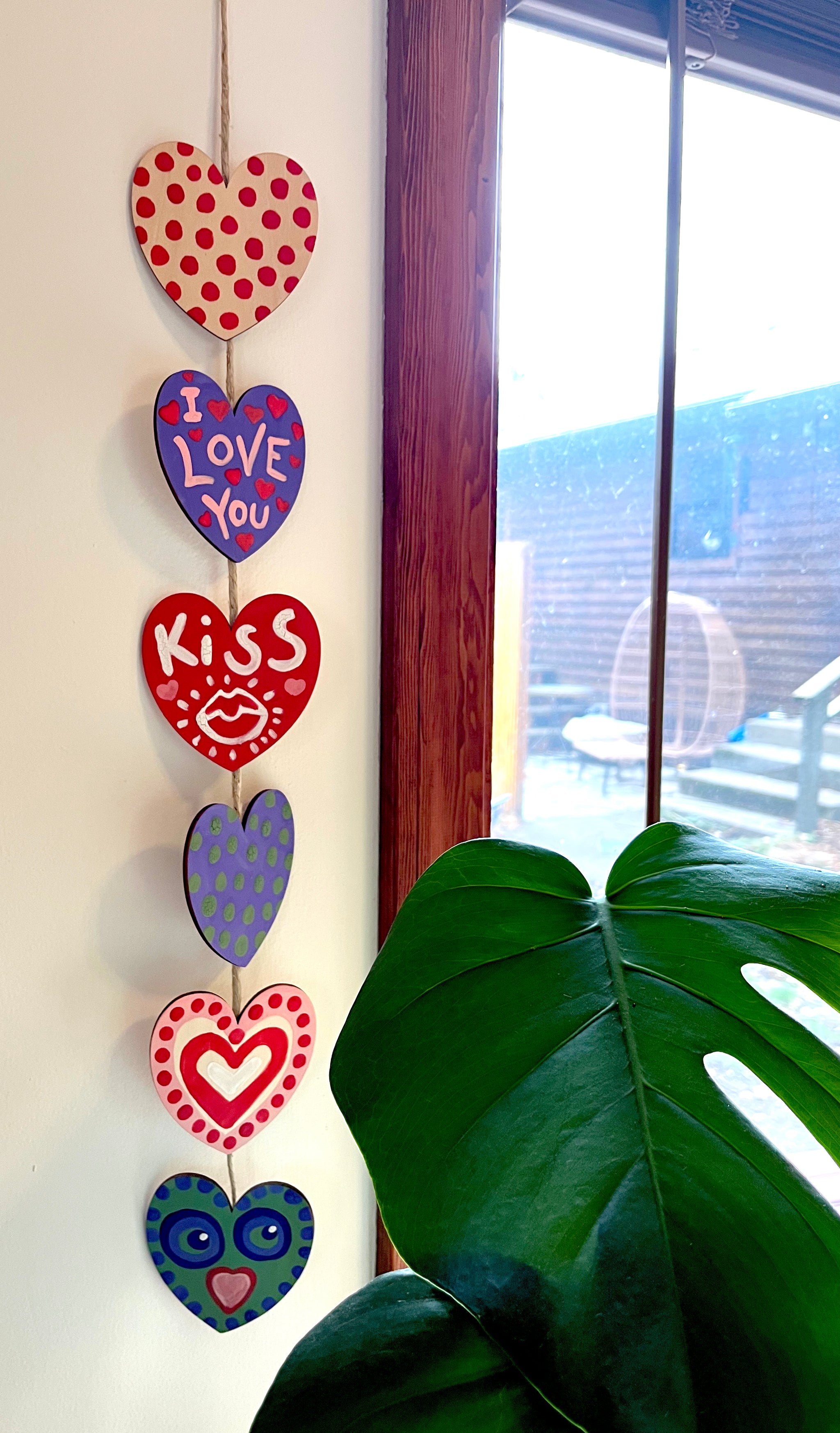 Image of 6 painted hearts attached by twine hanging on a all near a plant.