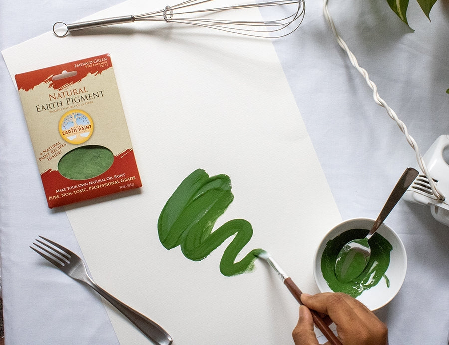 Person painting a green squiggle on a white piece of paper sitting next to a metal fork, green packet of earth & mineral pigment, metal whisk and bowl of green paint with a metal spoon resting in it