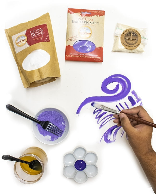 person's hand holding a paint brush painting purple gouache paint onto paper surrounded by the ingredients needed to make the paint