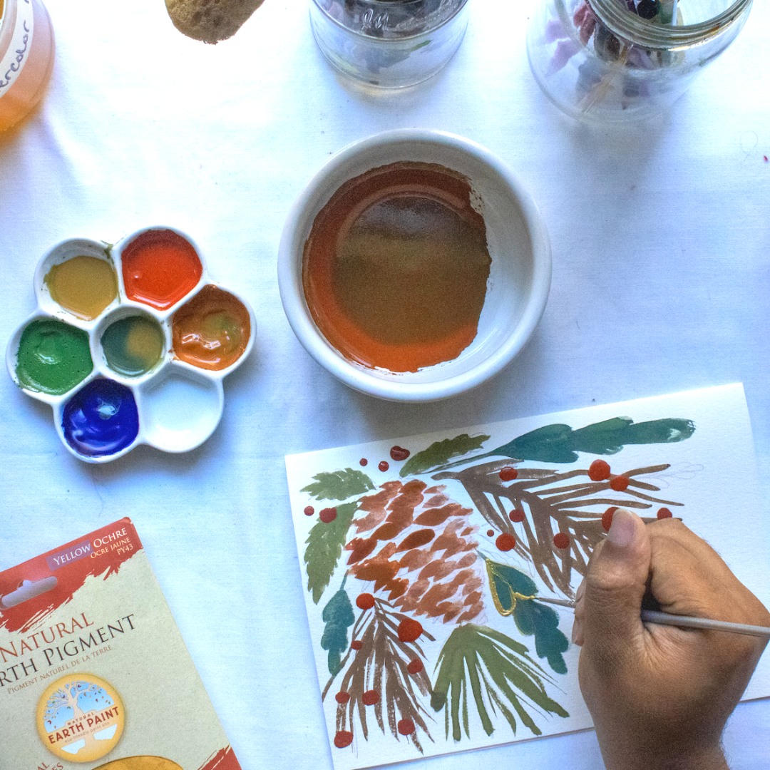 Person's hand holding a paint brush painting a white card with a pine cone and leaves next to a paint pallet and bowl with paint