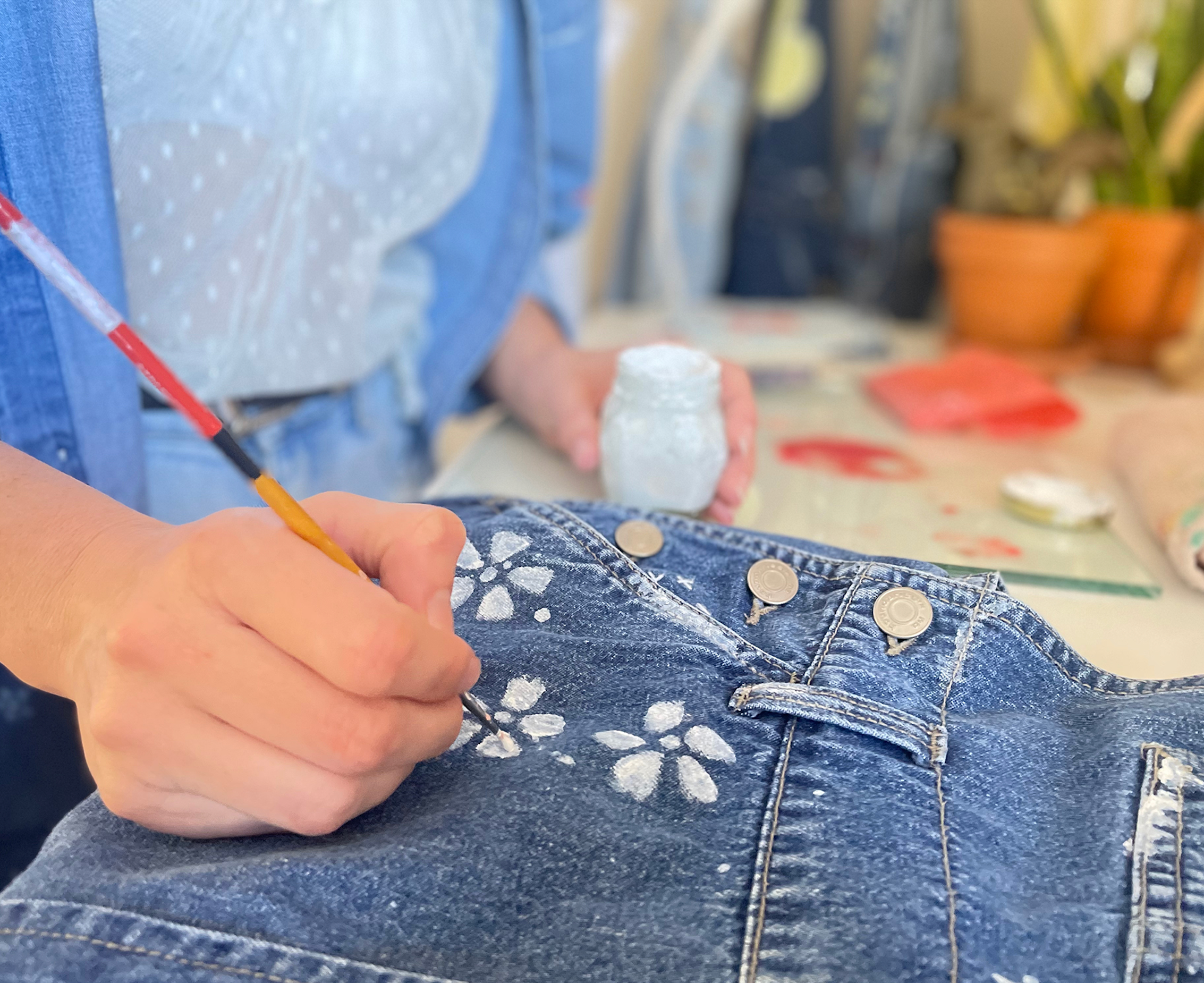 Close up of Artist Micaela Rish's hand painting white flowers on blue jean fabric using a paint brush