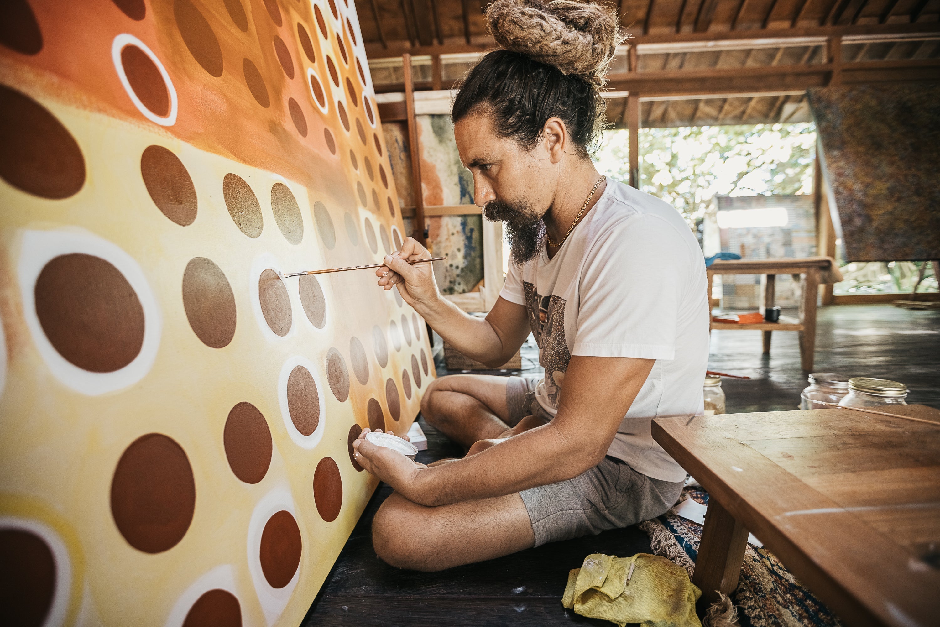 Portrait of artist Kristyan Stjerne sitting on the ground, working with a paint brush on a large painting in their studio 