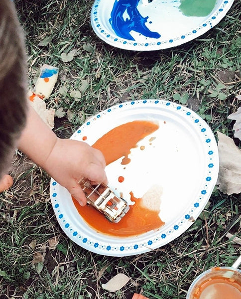 Child pressing a toy car into a pile of orange paint on a paper plate