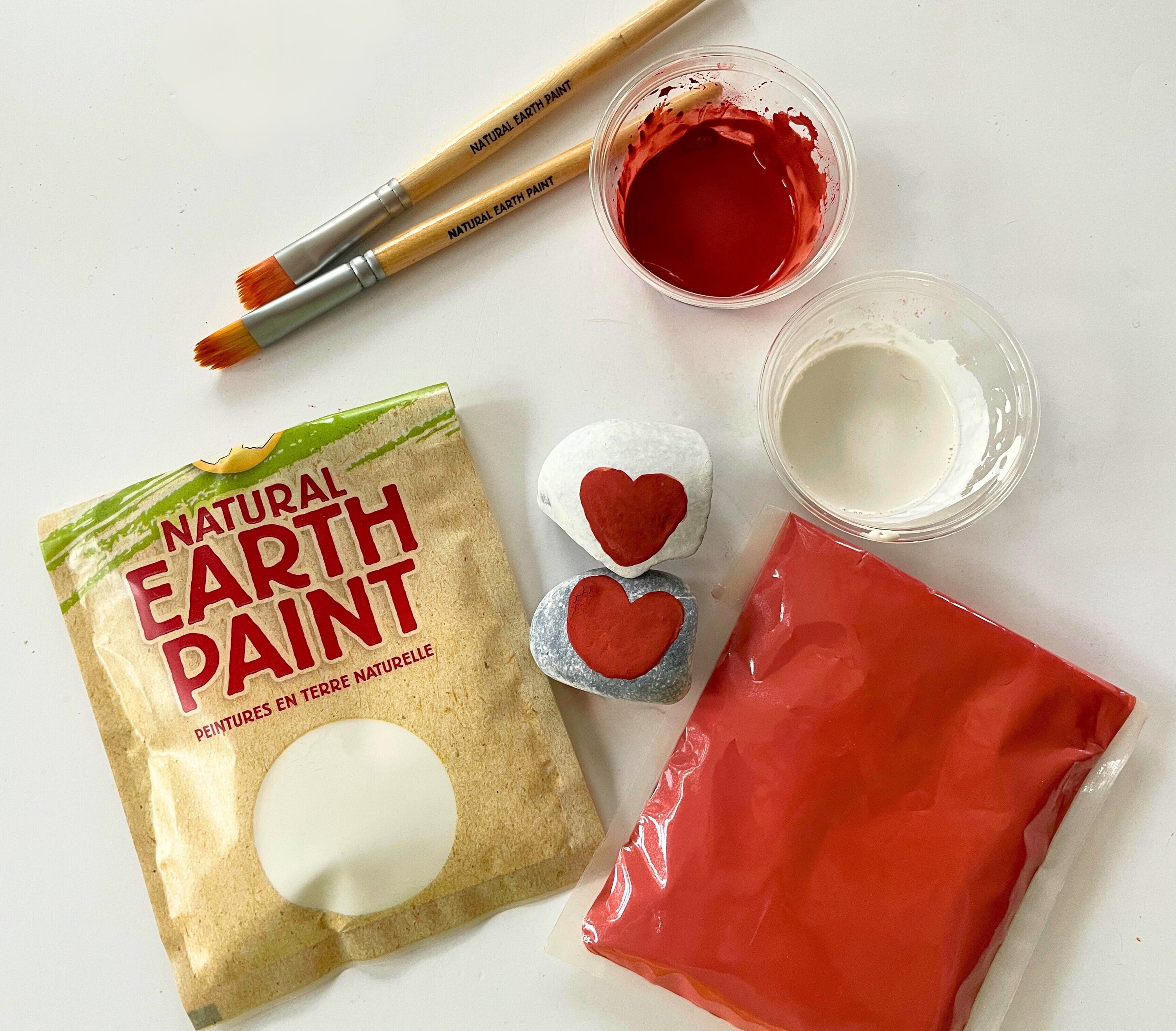 Natural Earth Paint pouches (red and white) with cups of paint, bamboo brushes, and two rocks painted with a red heart.