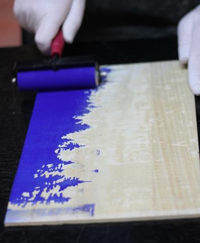 Person holding a brayer with blue ink, inking up a wood block with a tree design carved into it.