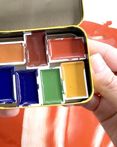 Person holding an aluminum tin filled with several small pans of handmade watercolor in various colors. 