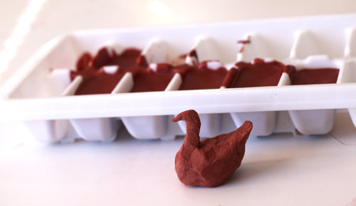 red shape clay bird sitting in front of ice cube tray full of red clay