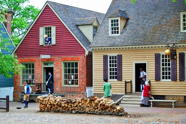 Scene of two old Colonial style buildings next to each other with a pile of firewood our front with Colonial dressed people 