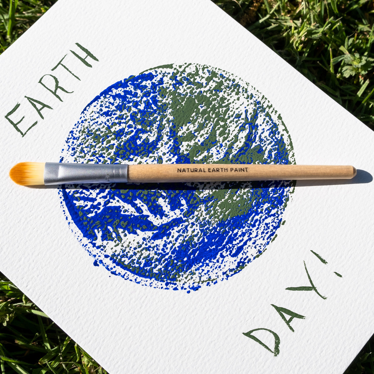 A painted card celebrating Earth Day, with a branded paint brush, on a patch of lawn