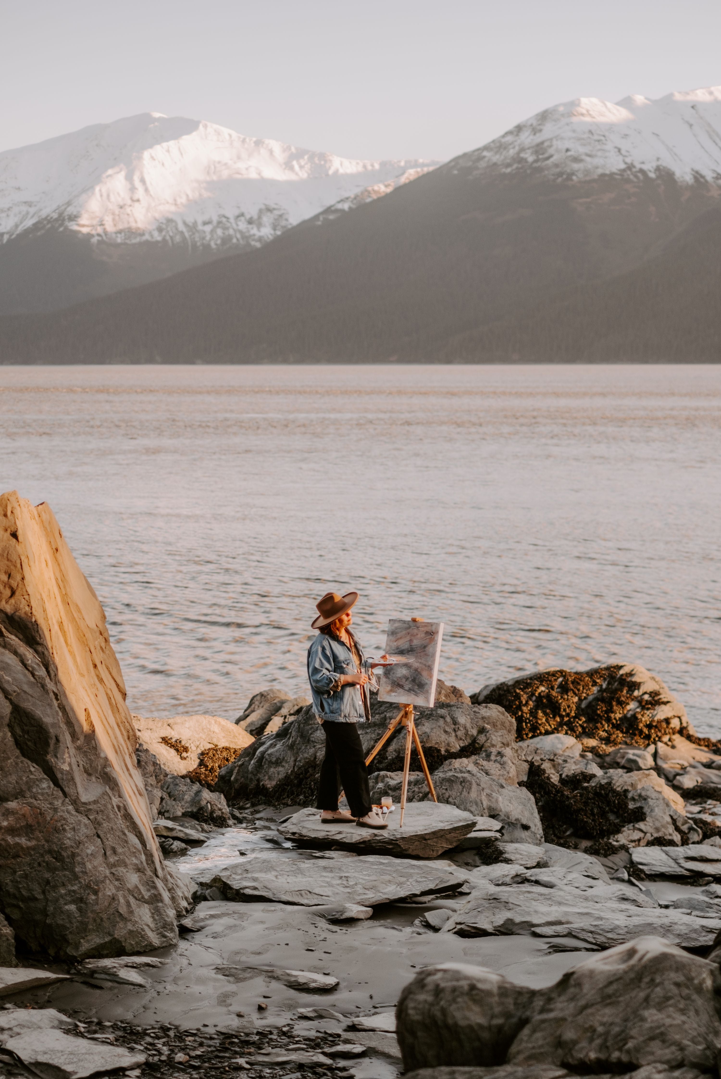 woman painting on canvas by water and mountains