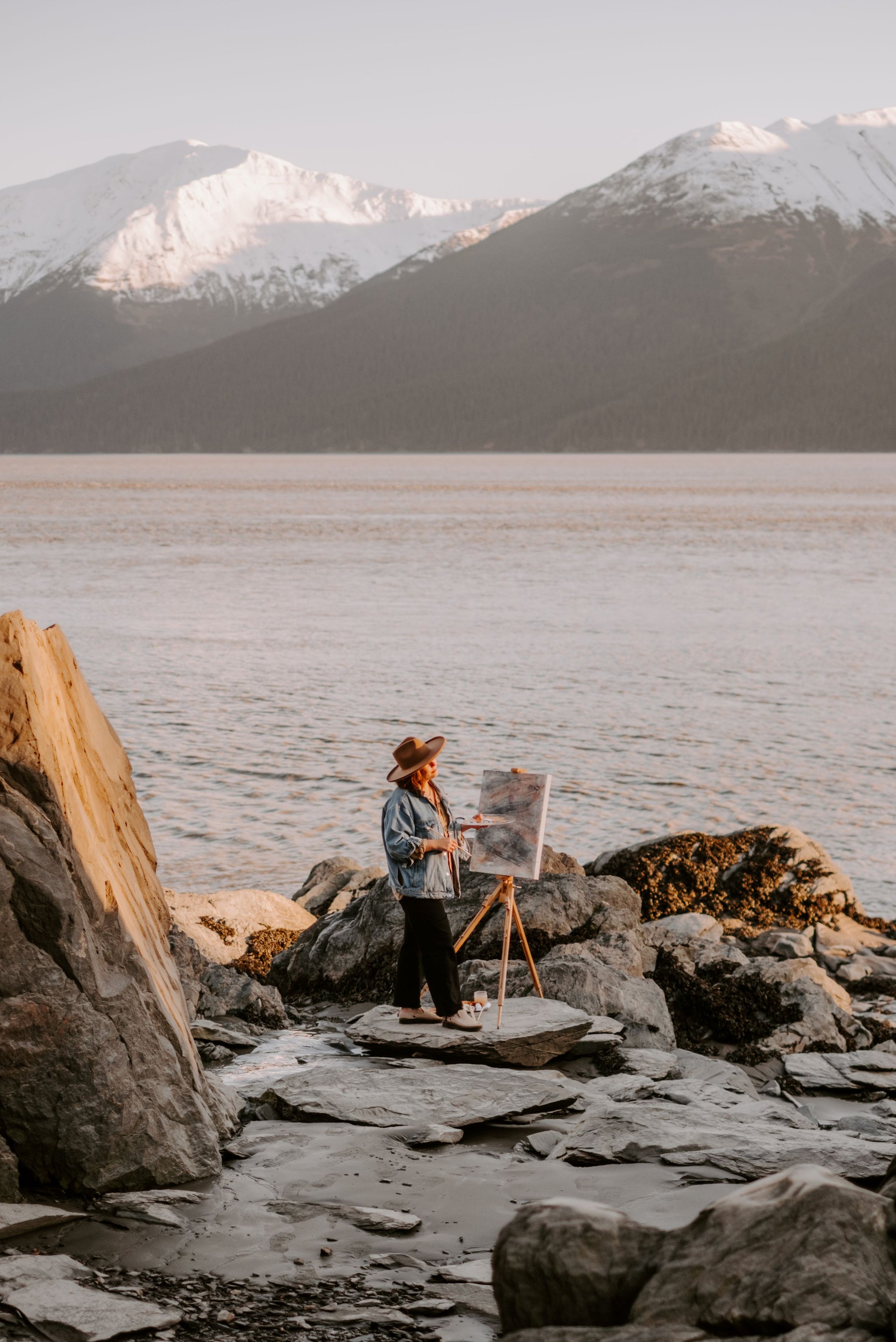 woman painting on canvas by water and mountains