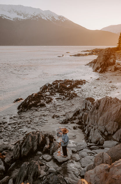 woman painting on canvas on a beach near mountains