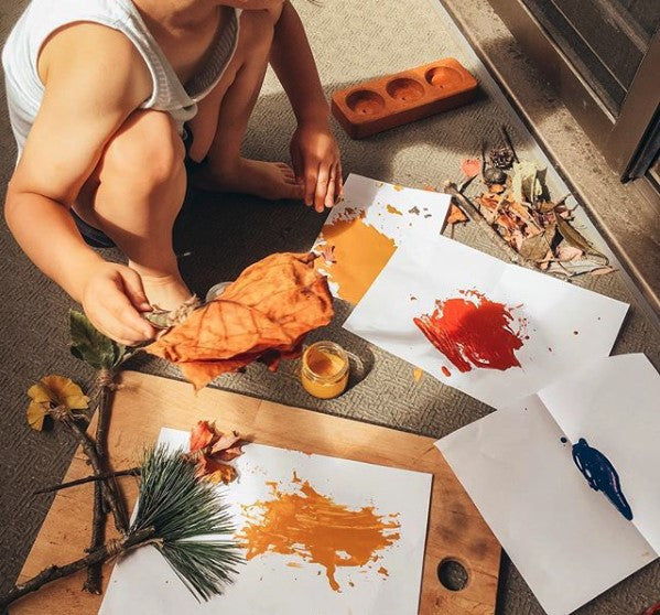 A small child painting warm colors on paper in the late afternoon with associated art supplies and samples of autumnal vegetation 