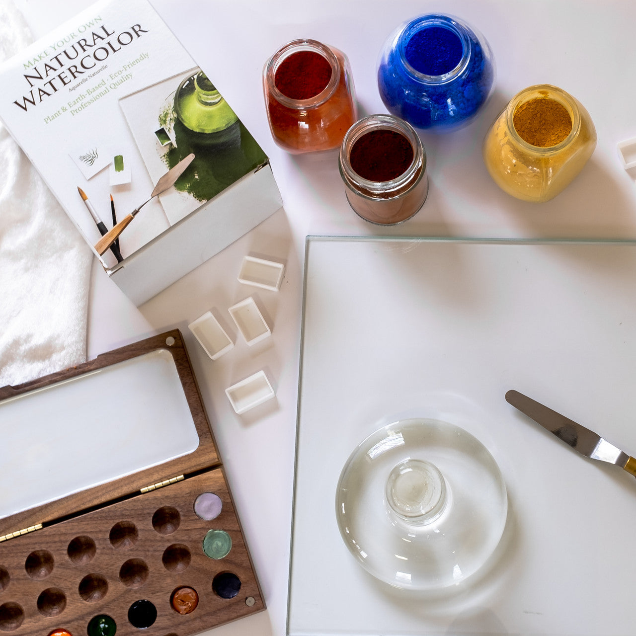 Watercolor supplies including glass palette, palette knife, glass muller, four open jars of paint, watercolor pans and a wooden watercolor palette