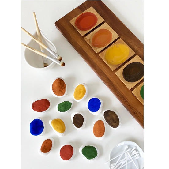 Wooden board supporting small trays of Natural Earth Paint alongside three paint brushes, some Q-tips and an array of stones painted in a minimalist style