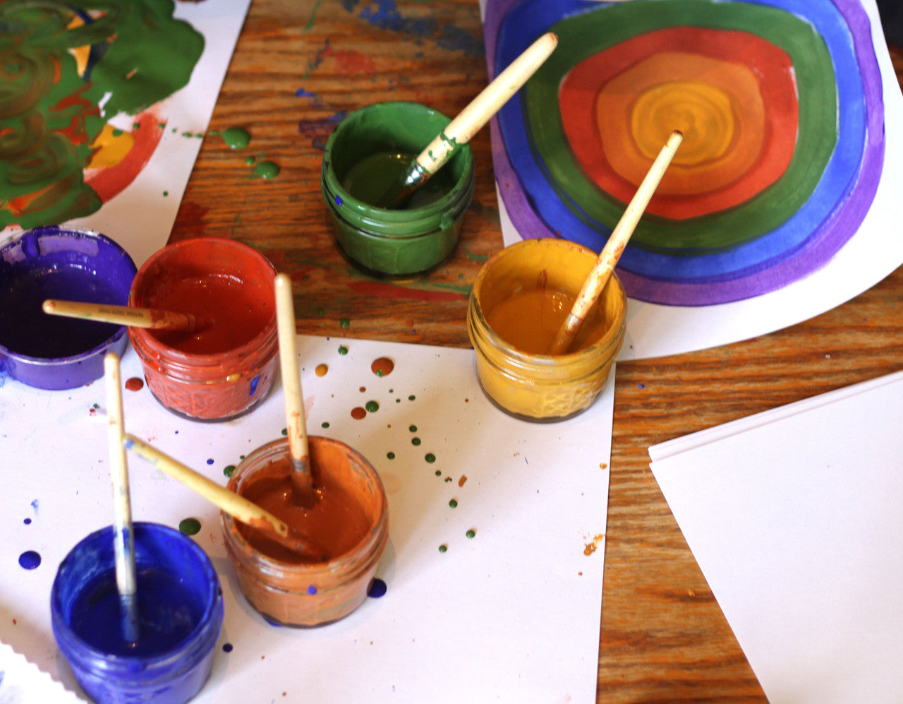 Six open jars of Natural Earth Paint with brushes and examples of children's art