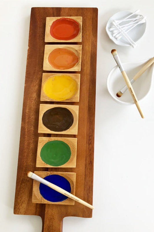 Wooden board with six colors of mixed paint shown with three paint brushes and some Q-tips